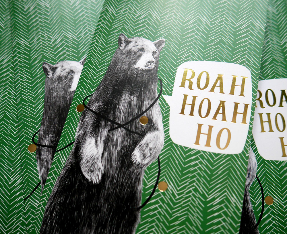 Photo of a Christmas card with two of the same card partially visible underneath. The cards have a green background with a white hand-drawn herringbone pattern. A bear tangled in Christmas baubles says 'Roah Hoah Ho'. The text and baubles are highlighted with gold foil.